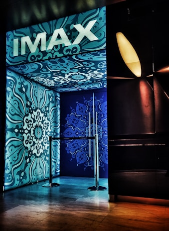 An entrance with ornate blue and teal designs covers the walls and ceiling, featuring intricate floral patterns. The brand 'IMAX' is prominently displayed at the top in bold white letters. A metallic barrier is present at the entryway, and ambient lighting creates a modern cinematic ambiance.