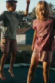 Two children are energetically bouncing on a trampoline. They are dressed in casual summer attire with the boy wearing a blue shirt and shorts, while the girl is in a pink top and shorts. The trampoline is situated outdoors, with trees and a building visible in the background, giving a sense of a backyard setting.
