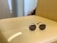 A pair of round sunglasses with dark lenses and thin metallic frames resting on a light tan leather surface. Soft ambient light casts a gentle reflection on the surface, creating a calm and serene setting. In the background, there is an out-of-focus section of a room, adding depth to the composition.