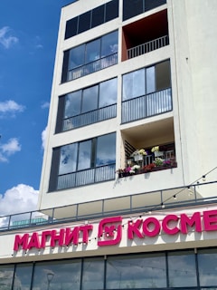 A modern building with large glass windows and a balcony decorated with colorful flowers. Below the balcony, there is signage with bold pink Cyrillic letters. The sky is clear with a few fluffy clouds.