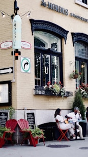A quaint outdoor caf&eacute; scene features a man and a woman seated at a small table with red chairs. The building's facade is cream-colored with black window trims and has flower boxes filled with red and white blooms. There are hanging signs, including a street direction sign, and a restaurant name 'Les Mordus' prominently displayed. A couple of red Adirondack chairs are placed nearby with potted plants.