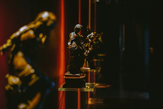 A sepia-toned photograph of a dimly lit gallery corner with textured paper walls and golden light highlighting a bronze sculpture.