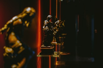 Bronze statues are displayed on glass pedestals in a dimly lit gallery, with dramatic red and dark shadows creating a mysterious and elegant atmosphere.