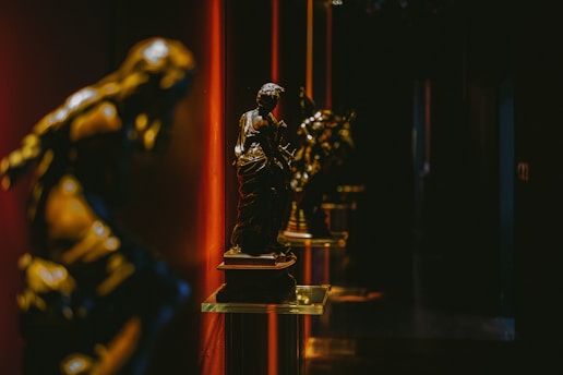 Bronze statues are displayed on glass pedestals in a dimly lit gallery, with dramatic red and dark shadows creating a mysterious and elegant atmosphere.