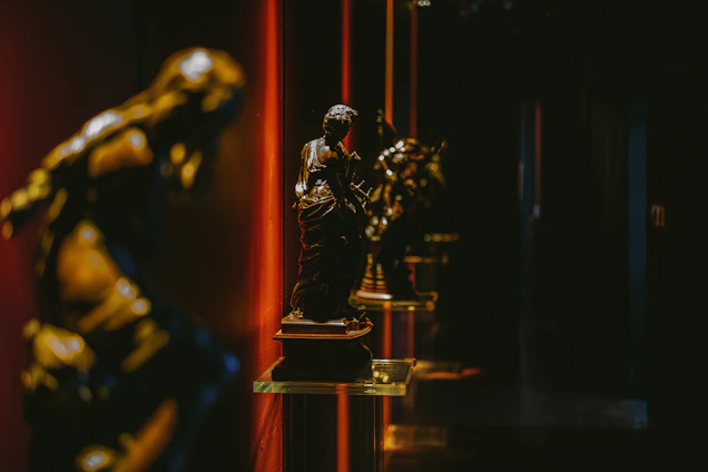 A sepia-toned photograph of a dimly lit gallery corner with textured paper walls and golden light highlighting a bronze sculpture.