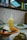 A dining table with a meal consisting of a yellow drink with a straw, a plate of rice topped with a fried egg and dark sauce, and a bowl of cooked green vegetables. The background features a wall with small square tiles and stained glass windows above.