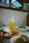 A dining table with a meal consisting of a yellow drink with a straw, a plate of rice topped with a fried egg and dark sauce, and a bowl of cooked green vegetables. The background features a wall with small square tiles and stained glass windows above.
