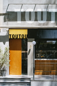 An urban storefront with a modern design features a large window and a wooden door. The building is made of light-colored stone, and there's a potted plant to the left of the entrance. A shadow of large text is cast across the facade, adding an artistic touch. A small lamp post stands near the entrance stairway, and part of the interior is visible through the window.