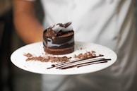A round chocolate dessert is beautifully presented on a white plate, garnished with chocolate shavings and decorated with artistic streaks of chocolate sauce. The dessert features layers of chocolate with a ribbon-like design on the top.