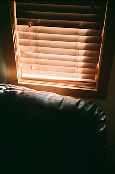 Wooden blinds partially opened with sunlight streaming through, casting shadows on a dark leather couch positioned below the window.