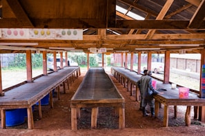 An open-air wooden structure with several long tables lined up, possibly used for drying or sorting agricultural produce. There are two people standing at the nearest table, engaged in handling some materials. The roof is supported by wooden beams, and there are vehicles parked outside along a fence in the background.