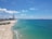 A scenic coastal view with a sandy beach stretching along the shoreline. On the left, there is an industrial area with tall smokestacks emitting white smoke. The ocean extends to the horizon, with clear blue skies and scattered clouds above.