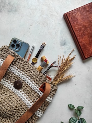 A woven bag with leather straps contains various items such as a smartphone, a pencil, a watch, lipstick, and dried wheat stalks. The bag rests on a light textured surface, accompanied by a closed brown leather journal and some small green leaves.