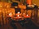 Several lit candles in metallic holders are being held in a dimly lit, warm and serene environment. In the background, muted gold and brown tones dominate the decor, with additional candles and religious or ceremonial objects placed on a table covered with a draped cloth.