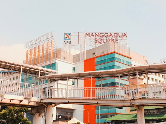 A large building complex features modern architecture with a prominent sign for Mangga Dua Square on top. The structure is designed with glass windows and an orange accent wall. An elevated pedestrian walkway crosses in front of the buildings.