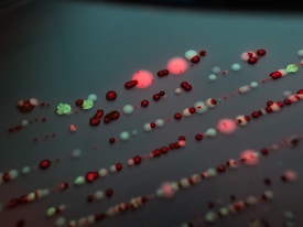 Various colorful colonies, including small red and green dots and larger clusters, are scattered across a dark surface in a pattern typical of microbial growth. The image has a detailed focus on the intricate arrangement and subtle textures of the colonies.