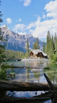 A picturesque lakeside scene featuring a rustic wooden cabin surrounded by pine trees with majestic mountains in the background. The clear blue sky is dotted with fluffy white clouds. A serene body of water reflects the cabin and surrounding nature, creating a tranquil atmosphere.