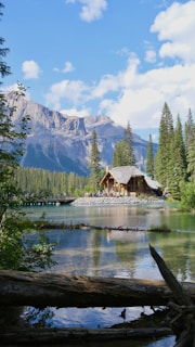 A picturesque lakeside scene featuring a rustic wooden cabin surrounded by pine trees with majestic mountains in the background. The clear blue sky is dotted with fluffy white clouds. A serene body of water reflects the cabin and surrounding nature, creating a tranquil atmosphere.