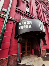 A red brick building with a sign in Russian above the entrance, featuring an illustrated face. The door is wooden and surrounded by darker red bricks. A green vertical pipe is visible on the left side.