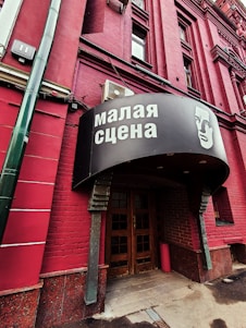 A red brick building with a sign in Russian above the entrance, featuring an illustrated face. The door is wooden and surrounded by darker red bricks. A green vertical pipe is visible on the left side.