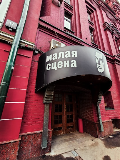 A red brick building with a sign in Russian above the entrance, featuring an illustrated face. The door is wooden and surrounded by darker red bricks. A green vertical pipe is visible on the left side.