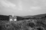 A small, white church with a steeple is situated in an expansive field of tall grass and wildflowers. It stands against a backdrop of rolling hills under a cloudy sky.