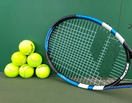 A set of bright yellow tennis balls is stacked in a pyramid alongside a tennis racket with a blue and white frame on a green court surface.