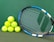 A set of bright yellow tennis balls is stacked in a pyramid alongside a tennis racket with a blue and white frame on a green court surface.