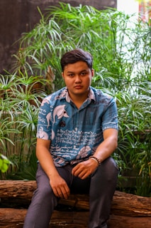 A person is sitting on a wooden log, surrounded by lush green plants. They are wearing a blue shirt with white and black graphics, and dark pants. Their expression is neutral, and they are seated with hands resting on their lap. The setting has an indoor garden feel with a focus on natural elements.