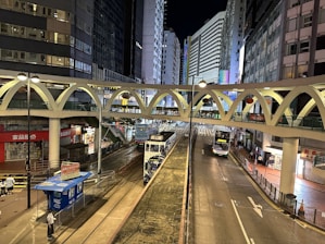 A nighttime urban scene featuring a busy street between tall buildings with a tram and a bus traveling in different lanes. An elevated pedestrian bridge with a distinct arc design crosses over the street. Bright city lights illuminate the scene, with various signs and shop logos visible at street level.