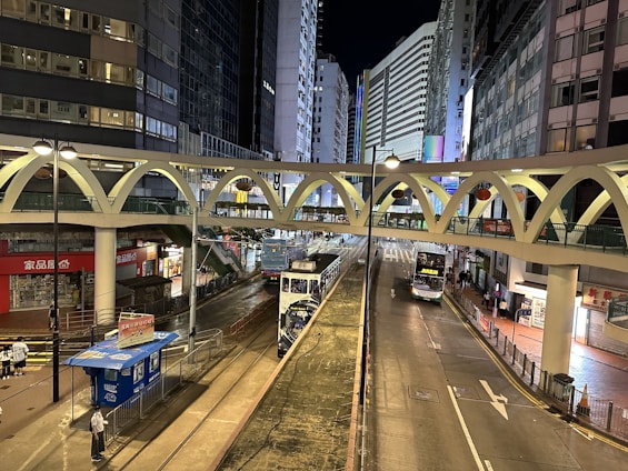 A nighttime urban scene featuring a busy street between tall buildings with a tram and a bus traveling in different lanes. An elevated pedestrian bridge with a distinct arc design crosses over the street. Bright city lights illuminate the scene, with various signs and shop logos visible at street level.