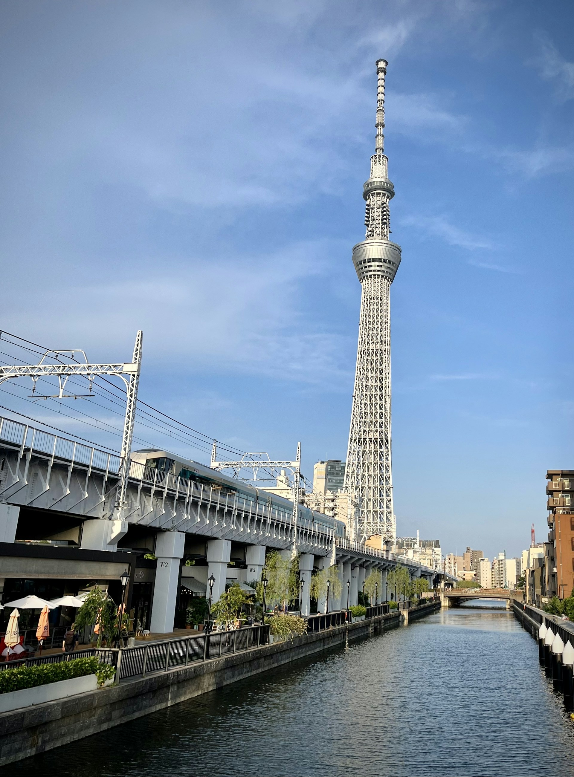 A tall broadcast tower stands against a clear blue sky, alongside a railway track and a canal bordered by urban buildings. The scene is set in a busy city environment with clear weather.