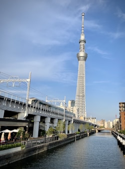 A tall broadcast tower stands against a clear blue sky, alongside a railway track and a canal bordered by urban buildings. The scene is set in a busy city environment with clear weather.