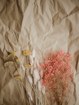 Several types of dried flowers are artistically arranged on a crumpled brown paper background. Yellow flowers are on the left, white fluffy stalks are in the center, and a bunch of small pink flowers is on the right.