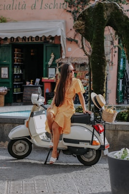 A woman in a yellow dress sits on a white scooter parked in front of a quaint Italian storefront. She wears sunglasses and is surrounded by a cobblestone street and greenery. A straw hat and basket are attached to the scooter.