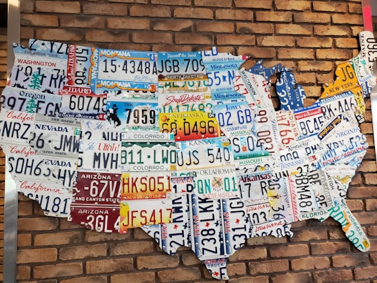 A collection of various U.S. license plates arranged to form the shape of the United States against a red brick wall. Each license plate represents a different state, showcasing a variety of colors and state-specific slogans or symbols.