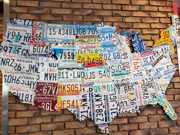 A collection of various U.S. license plates arranged to form the shape of the United States against a red brick wall. Each license plate represents a different state, showcasing a variety of colors and state-specific slogans or symbols.