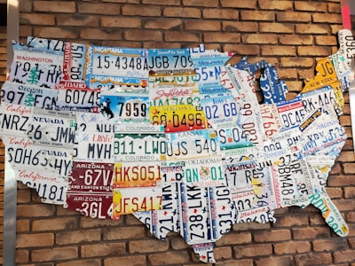 A collection of various U.S. license plates arranged to form the shape of the United States against a red brick wall. Each license plate represents a different state, showcasing a variety of colors and state-specific slogans or symbols.