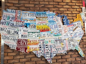 A collection of various U.S. license plates arranged to form the shape of the United States against a red brick wall. Each license plate represents a different state, showcasing a variety of colors and state-specific slogans or symbols.