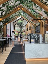 A modern, indoor cafe or ice cream shop with a high ceiling adorned with wooden beams and green plants. The counter features a sign labeled 'Gelato' with a display of desserts or ice cream containers. Several people are casually dressed, standing near the counter and the seating area. Floor is tiled with a patterned design while bar-style seating with tall chairs is visible to the left.
