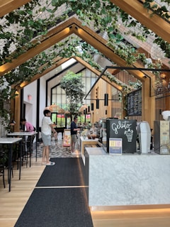 A modern, indoor cafe or ice cream shop with a high ceiling adorned with wooden beams and green plants. The counter features a sign labeled 'Gelato' with a display of desserts or ice cream containers. Several people are casually dressed, standing near the counter and the seating area. Floor is tiled with a patterned design while bar-style seating with tall chairs is visible to the left.