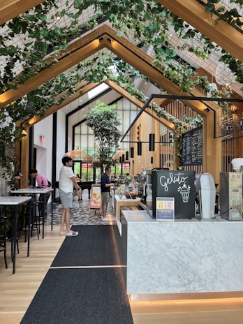 A modern, indoor cafe or ice cream shop with a high ceiling adorned with wooden beams and green plants. The counter features a sign labeled 'Gelato' with a display of desserts or ice cream containers. Several people are casually dressed, standing near the counter and the seating area. Floor is tiled with a patterned design while bar-style seating with tall chairs is visible to the left.