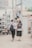 A boy and a girl wearing school uniforms and backpacks walk together on a city sidewalk. The boy gently places a hand on the girl's head while they stroll. The background features urban architecture with buildings, utility poles, and wires.