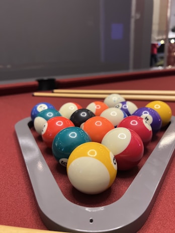 A neatly arranged set of pool balls sits within a triangular rack on a red felt pool table, accompanied by two cue sticks lying parallel to each other.