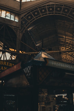 A dimly lit indoor market scene with a focus on intricate metalwork and a pastel shop sign. The structure features ornate iron arches and glass roofing. The pastel sign indicates a shop selling pastries, while another sign mentions specialty roasted coffee.