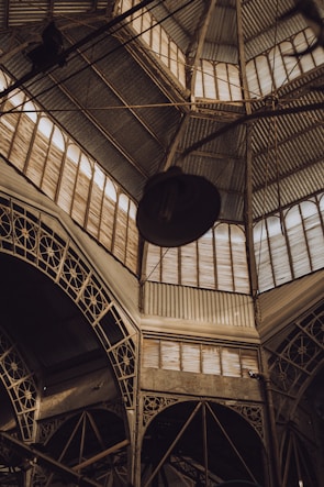 A geometric architectural structure featuring intricate metal framework and a corrugated metal roof. Light filters through translucent panels, casting shadows and highlighting the complex latticework.