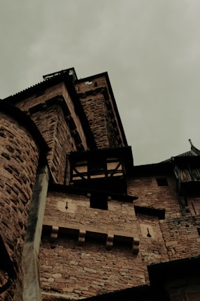 A medieval stone castle with dark, thick walls and towers viewed from a low angle, emphasizing its imposing and historic architecture. The sky is overcast, creating a somber atmosphere.