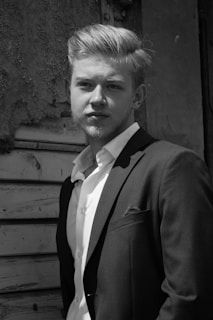 A man in a suit stands confidently in front of a textured, aged wooden background. The light highlights his facial features and well-groomed hair, giving a classic and timeless feel.