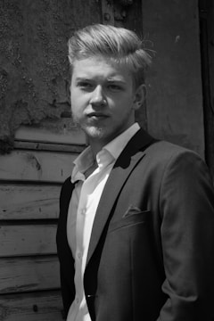 A man in a suit stands confidently in front of a textured, aged wooden background. The light highlights his facial features and well-groomed hair, giving a classic and timeless feel.