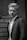 A man in a suit stands confidently in front of a textured, aged wooden background. The light highlights his facial features and well-groomed hair, giving a classic and timeless feel.